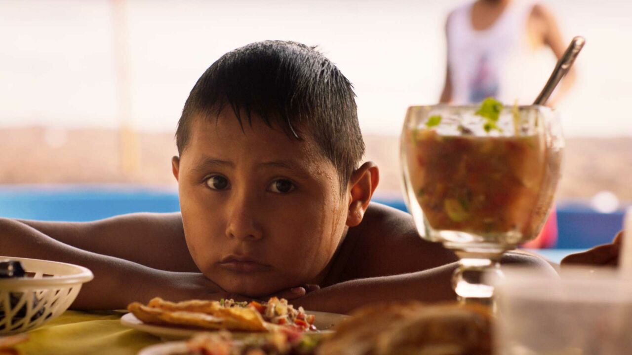 A child with medium skin tone and dark hair, looking downcast, sits with his arms and head resting on a table full of food.