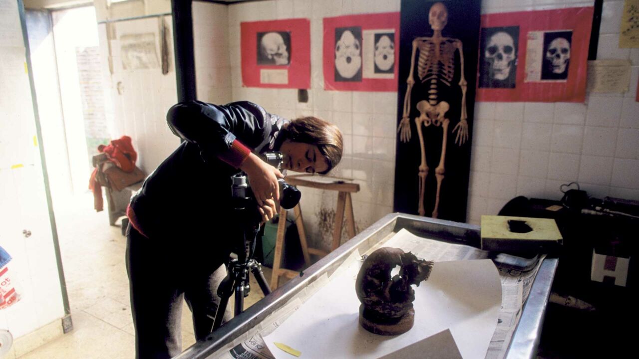 A woman with light skin tone, brown hair, and a leather jacket photographs a human skull in a forensic lab. Various photographs of human skulls and skeletons hang on the wall behind her.