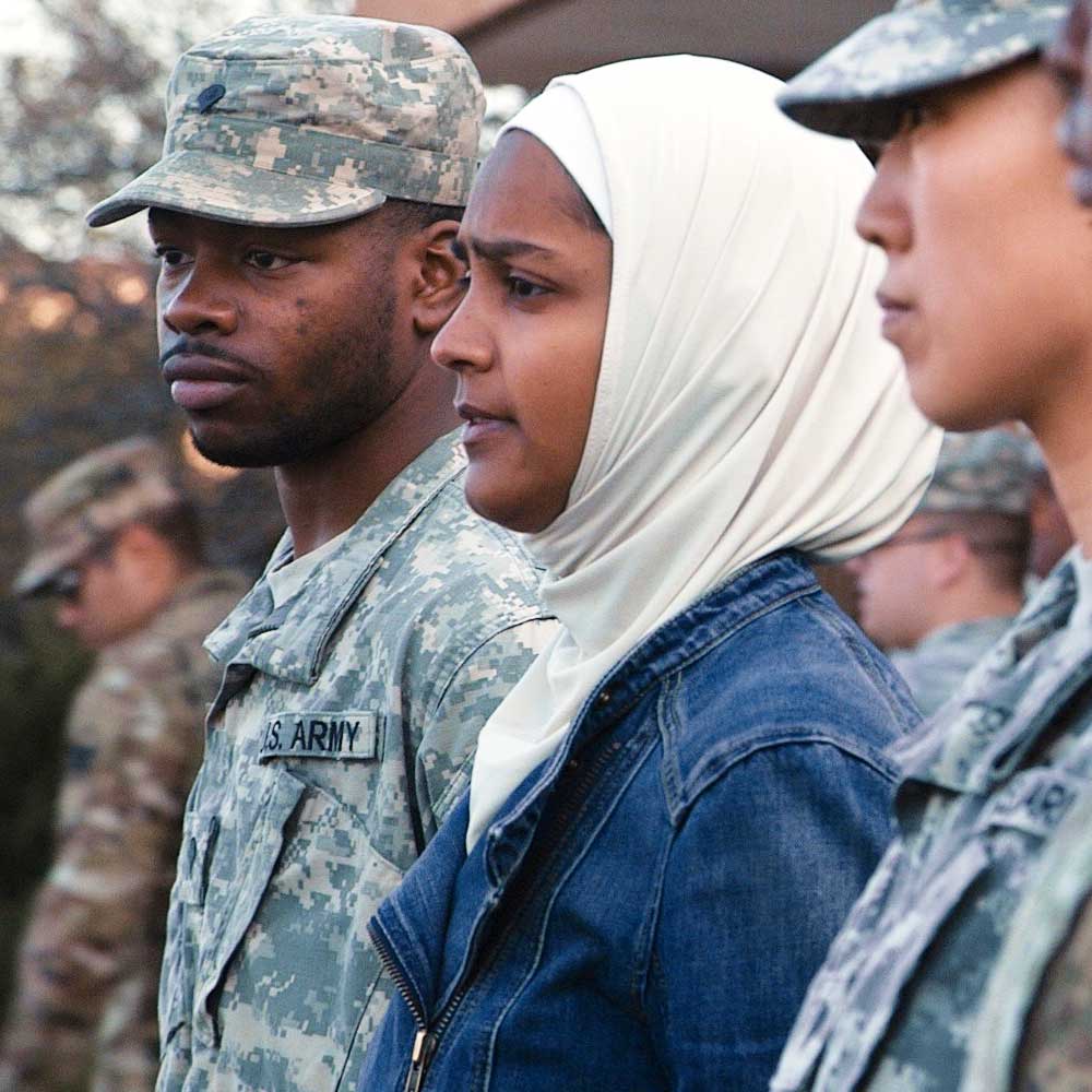 A man in a US military uniform with dark skin tone stands beside a woman with medium skin tone wearing a denim jacket and white hijab. Other soldiers stand in the background and foreground.