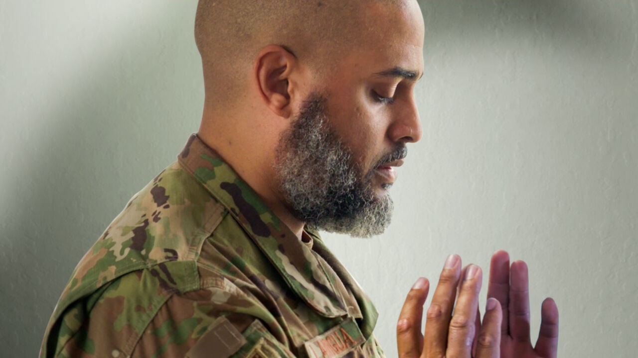 A man with medium skin tone, shaved head, and salt and pepper beard wears a US military uniform and bows his head in prayer.