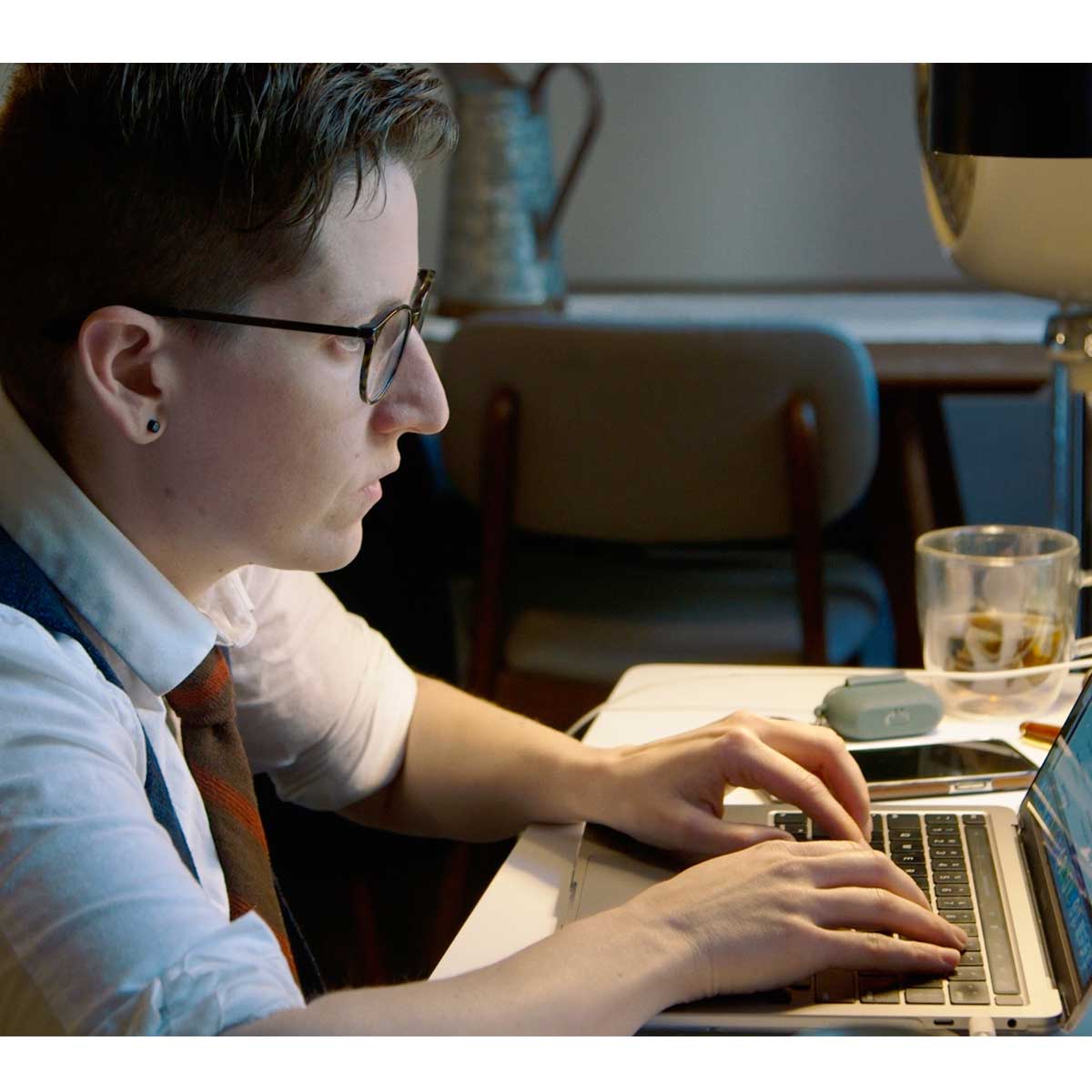 A person with light skin tone and short dark hair types on a laptop. They are wearing a vest and tie and look to be in deep concentration.