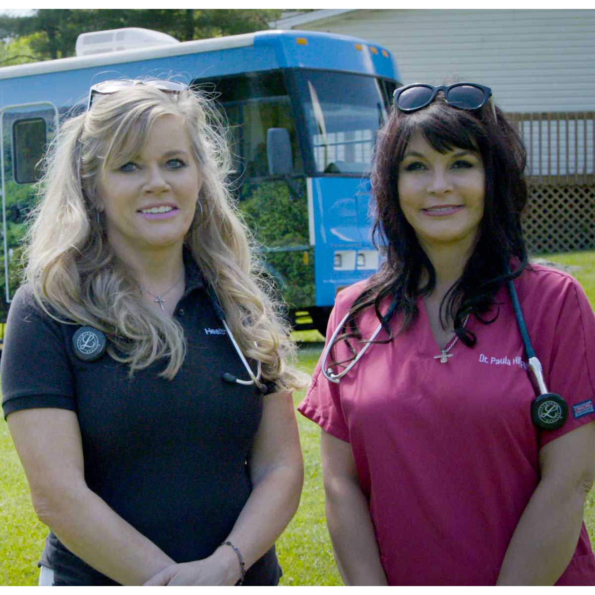 Two female doctors, both with light skin tone, pose outside of their mobile health wagon.