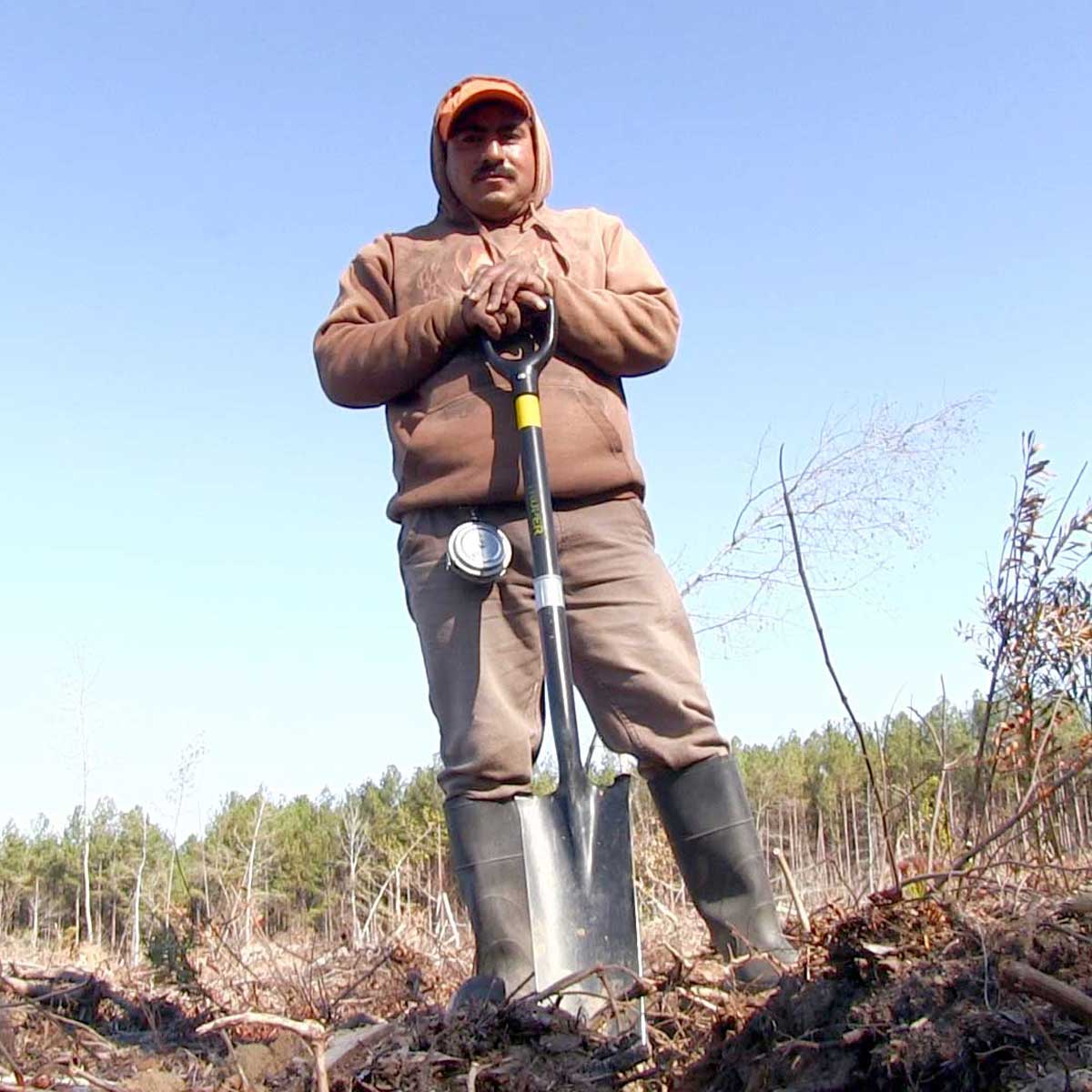 A man with medium-dark skin tone and a large black mustache poses with a shovel in a sparse forest. He wears a thick sweatshirt, hat, and knee-high boots.