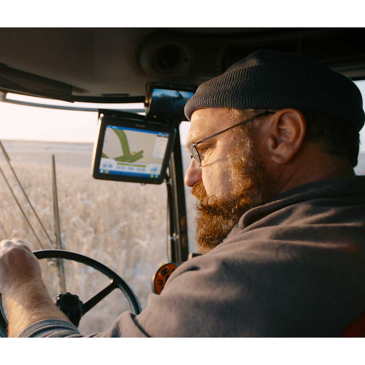 A man with light skin tone, a red beard, beanie, and glasses drives a tractor through a filed of corn that has been lightly dusted with snow.