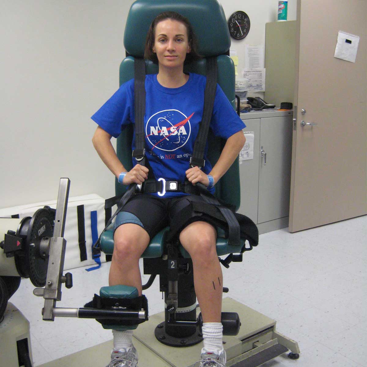 A woman with light skin tone and dark hair in a NASA tee shirt sits in a isokinetic testing chair in a laboratory.