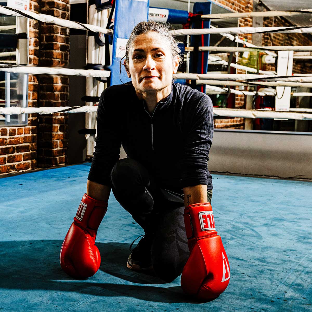 A woman with medium-light skin and dark hair wears boxing gloves while kneeling inside of a boxing ring.