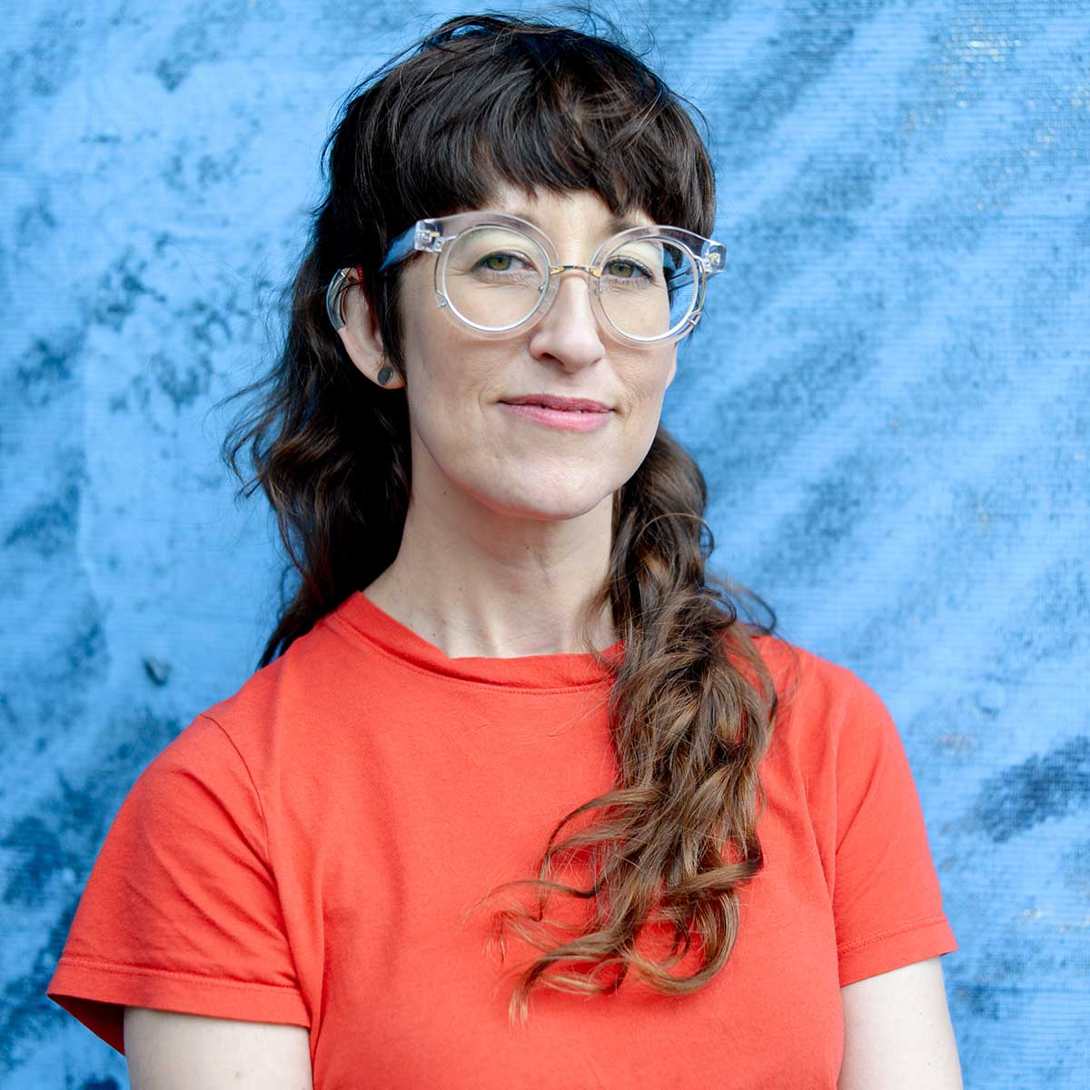 Headshot of Producer/Director Alison O'Daniel, a woman with light skin tone and chest-length brown hair. She wears clear-framed, round glasses and a bright orange shirt against a blue background.