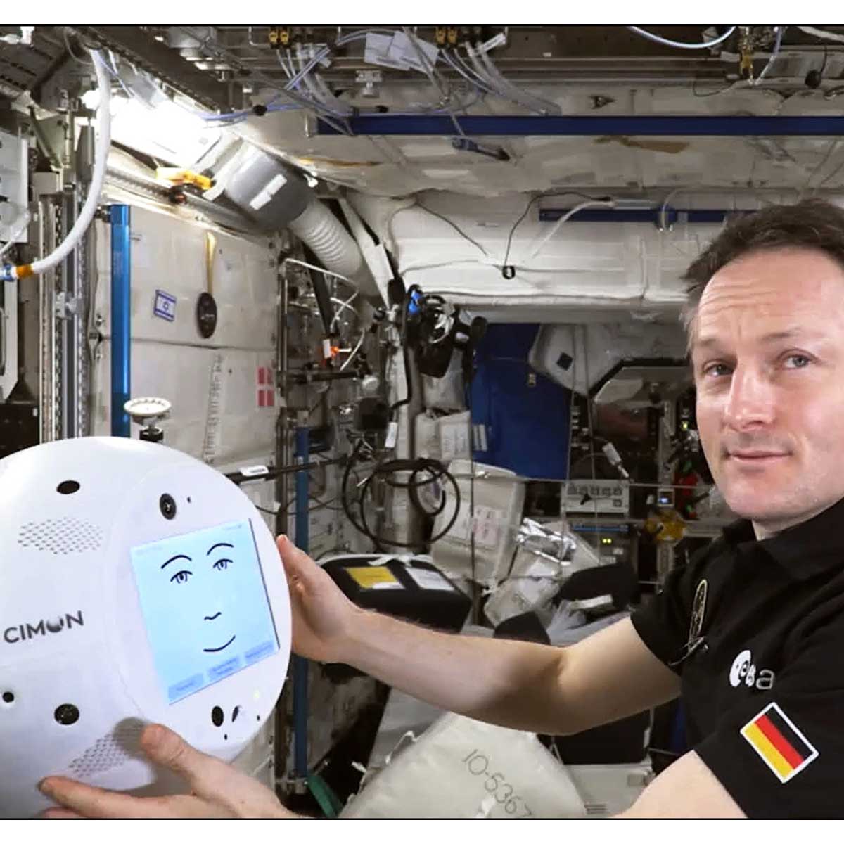 A German astronaut aboard a space station looks at the camera while holding CIMON, a robotic companion with a digital smiley face on its monitor.