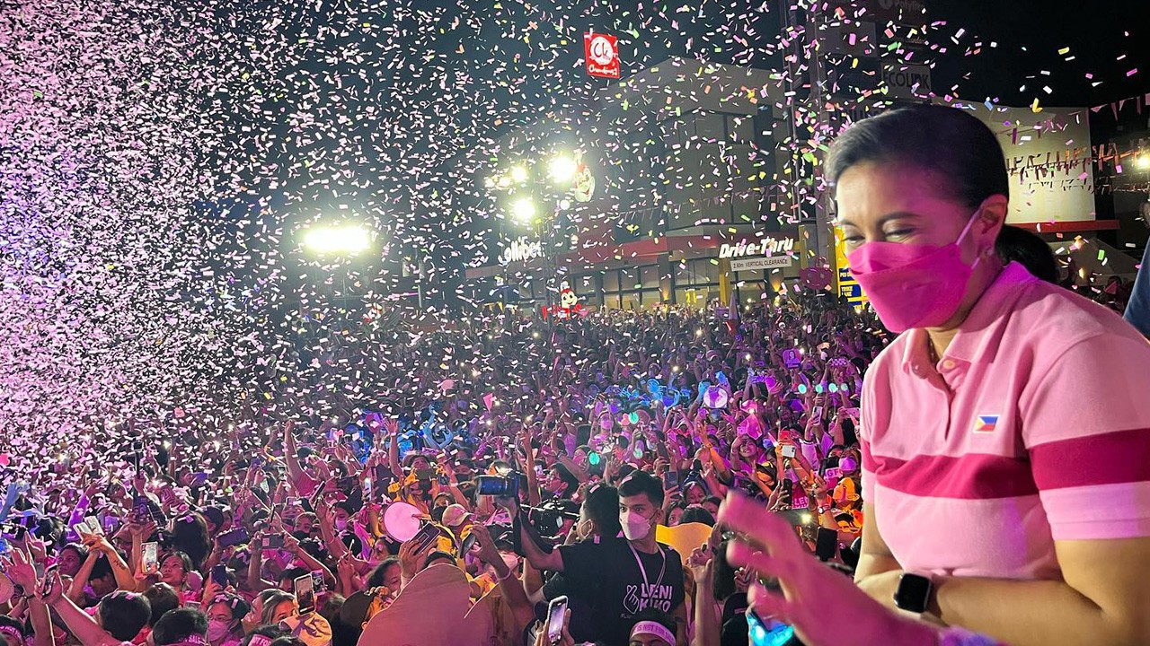 A woman outside a Jolibee restaurant in the Philippines wears a pink shirt and pink mask as she overlooks a crowd of people cheering under pink confetti.