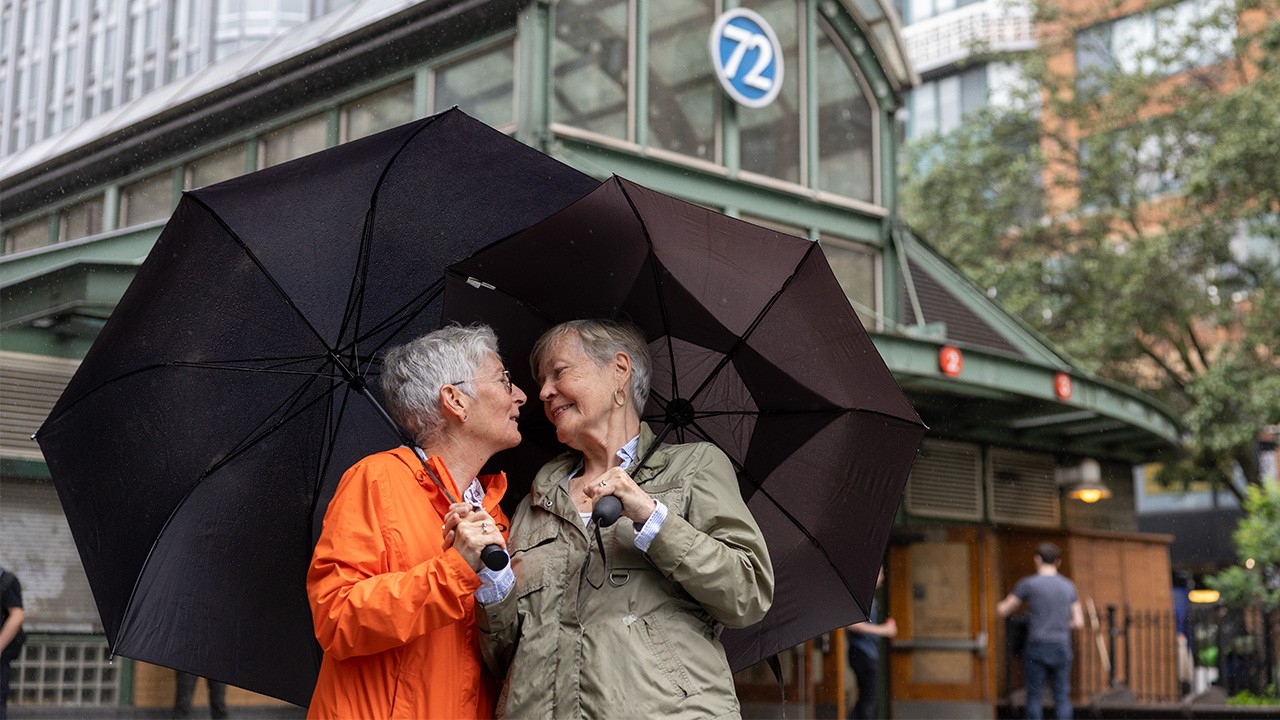 Two women with short gray hair look into each other's eyes outside in a city while holding umbrellas.