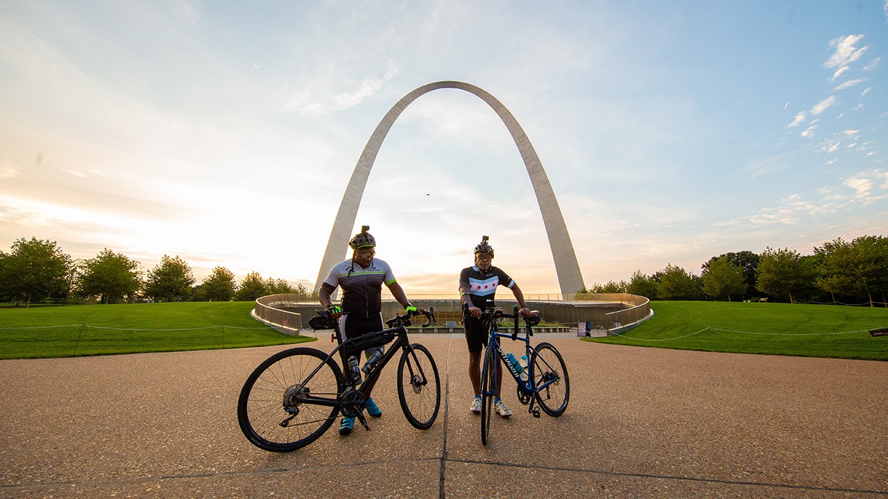 Two men with dark skin tone, a father and son, wearing professional bike gear and helmets while standing in front of the St. Louis Arch with their bicycles at sunset.