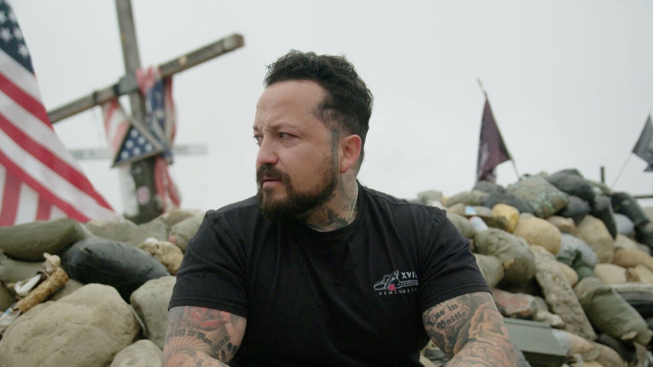 man with tattoos sitting in front of make shift wooden crosses with american flags on them