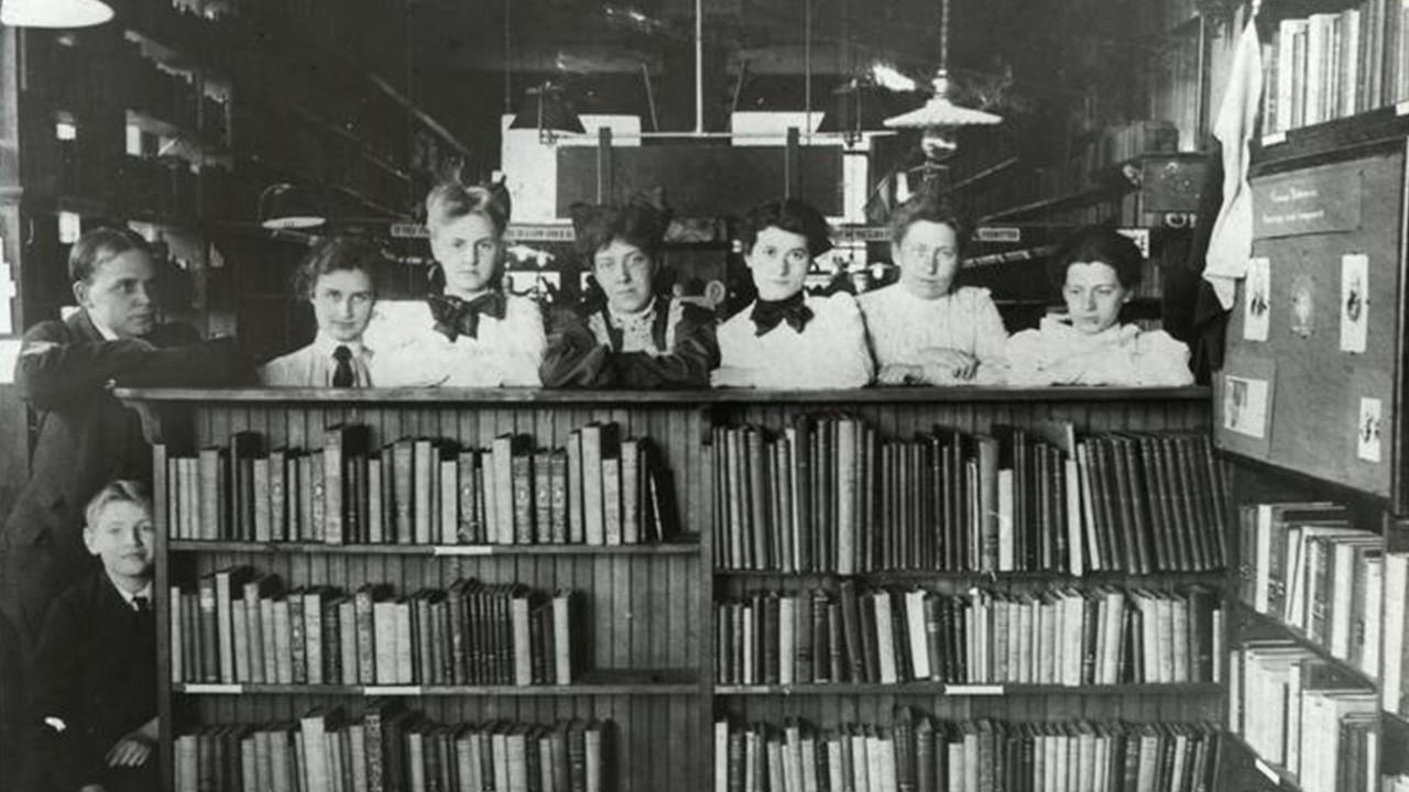 Black and white photograph of six women, one man, and a child in 1800s clothing standing behind a well-stocked bookshelf in a library.