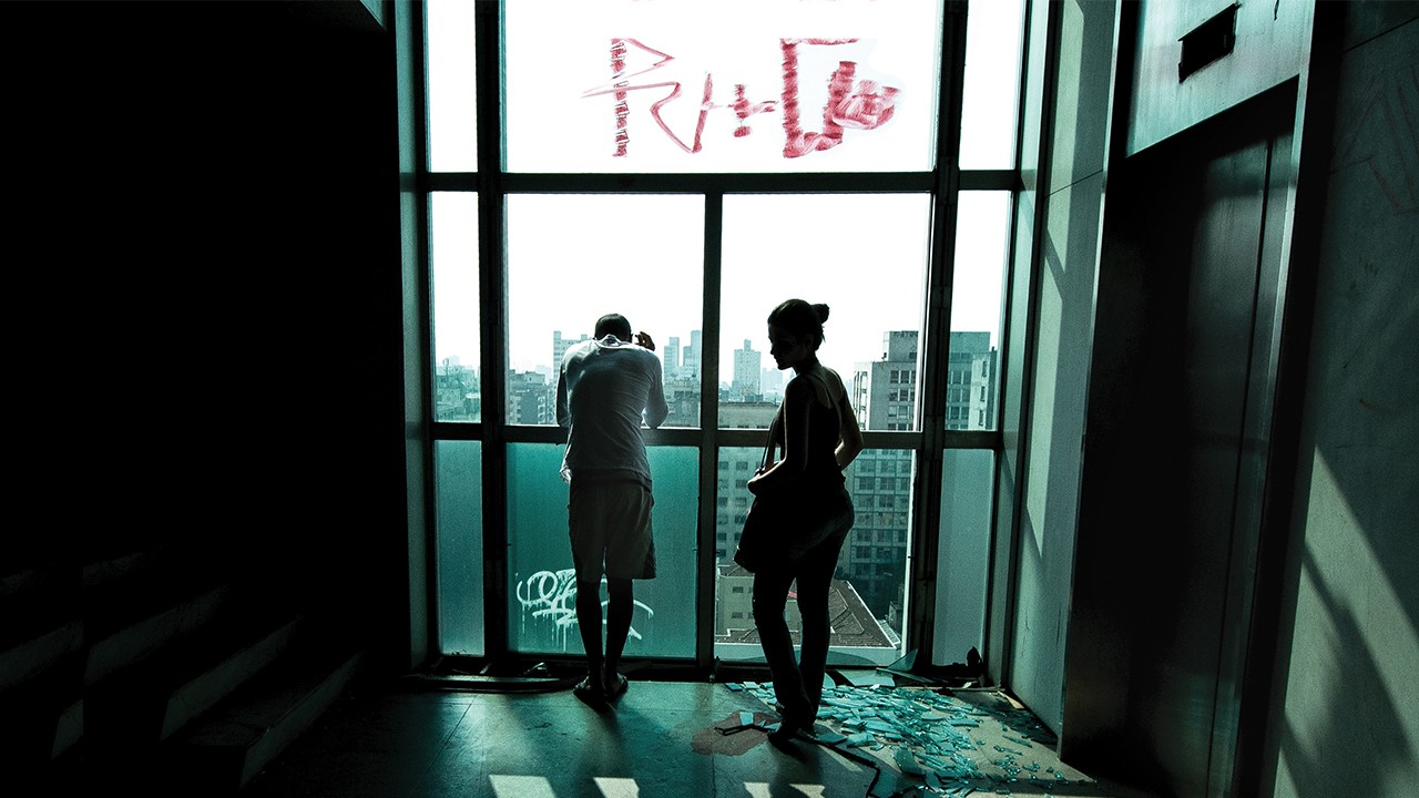 Photograph of a man and woman looking at a cityscape out of floor-to-ceiling windows covered in graffiti. One of the windows is broken and glass is scattered along the floor.